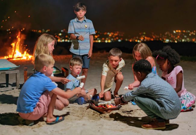 A Group Of People Sitting On The Sand By A Fire